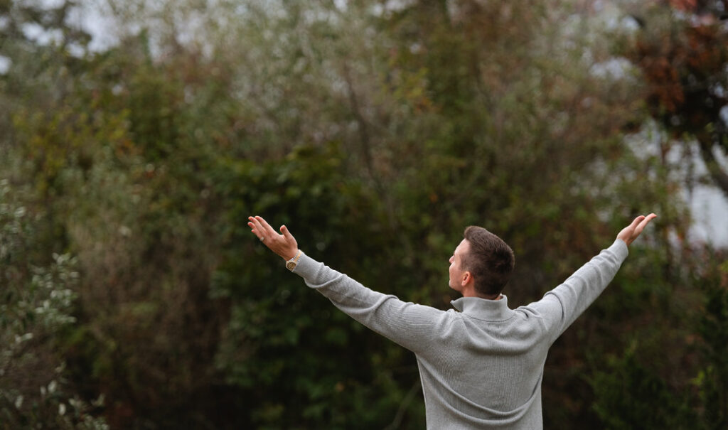 A person is standing outdoors with arms outstretched, facing a backdrop of dense greenery. Let's replace the mention of a man with the name Matt Fraser if applicable:

Matt Fraser is standing outdoors with arms outstretched, facing a backdrop of dense greenery.